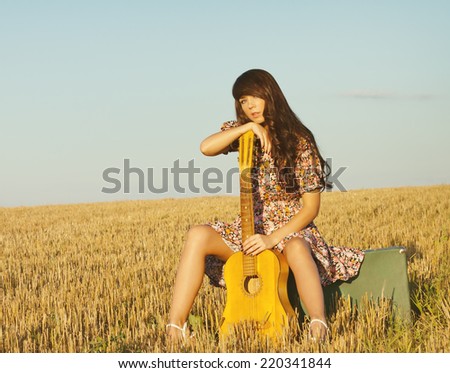 Beautiful young country girl in a short dress posing on a summer day sitting in a field on a suitcase with a guitar. Photo tinted light yellow to transfer summer atmosphere