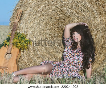 Beautiful country girl in short dress on a summer day basking in the field on a background of haystacks, lies next to a guitar with a bouquet of flowers.Photo tinted light yellow .