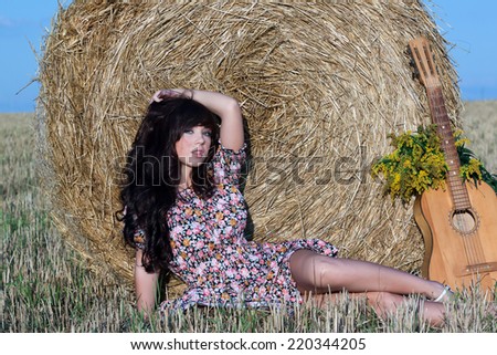 Beautiful country girl in short dress on a summer day basking in the field on a background of haystacks, lies next to a guitar with a bouquet of flowers.
