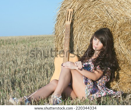Beautiful country girl in short dress on a summer day basking in the field on a background of haystacks, lies next to a guitar Photo tinted light yellow .