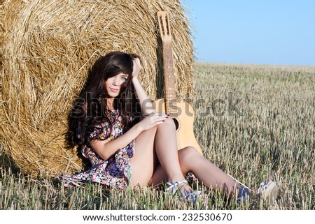 Beautiful country girl in short dress on a summer day basking in the field on a background of haystacks, lies next to a guitar Photo tinted light yellow .