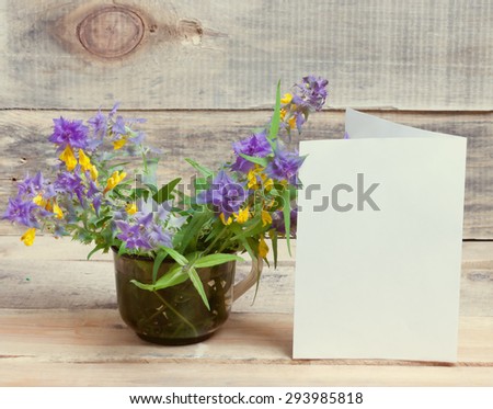 cup and a bouquet of wild flowers and a blank sheet of paper on a wooden surface.\
great blank for your design.closeup.toned