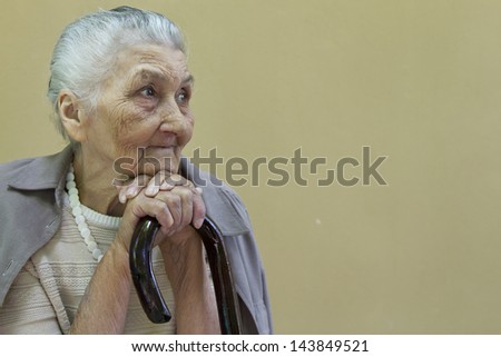 sad old lady\'s portrait with a walking stick  in front of a light brown wall