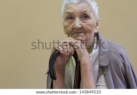 sad old lady\'s portrait with a walking stick  in front of a light brown wall