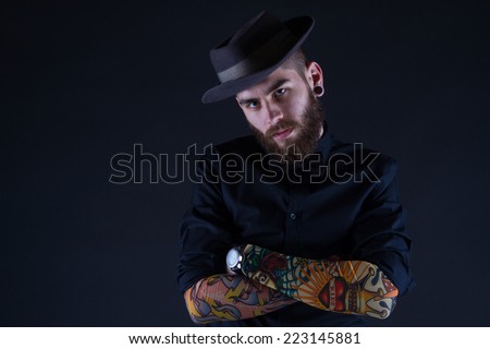 studio portarit of a fashionable  young  hipster man wearing stylish hat, tatooed sleeves and  posing over   a black background