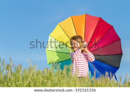 Girl stands in meadow with green grass against blue sky, watching sun and holding colored umbrella