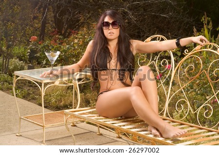 Young sensual woman in black bikini sitting on iron garden bench outdoors sunbathing, with cocktail glass on table by her side.