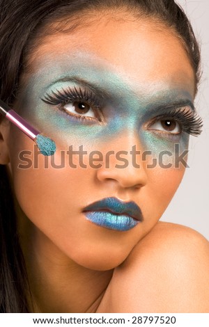 Young female beauty fashion model of mixed Creole and African-American ethnicity covered in dramatic blue make-up and holding makeup brush