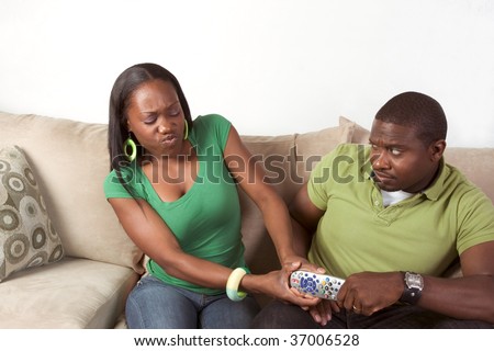Young black African American couple sitting in living room on couch and arguing over television remote control