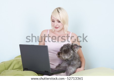 Caucasian blond young woman sitting on bed working on portable notebook pc computer together with dog of Schnauzer breed