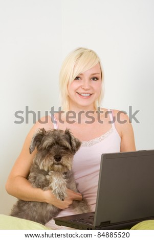 Caucasian blond young woman sitting on bed working on portable notebook pc computer together with dog of Schnauzer breed