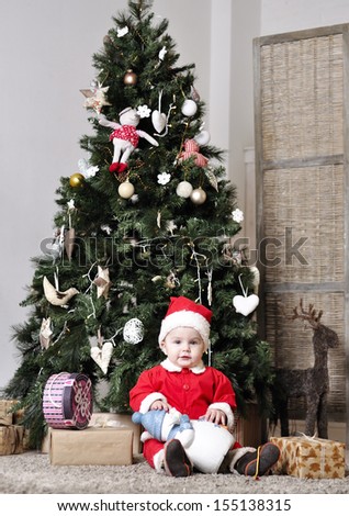 Baby in Santa costume sit near decorating Christmas tree with present toy