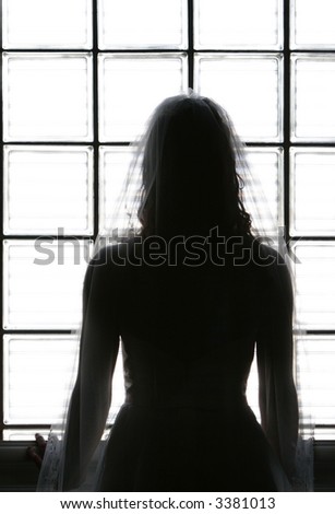 Back silhouette view of bride looking out a window with a pensive, anticipating feel, hands on window sill