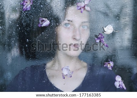 Closeup portrait of beautiful brunette woman behind the window with water drops on it and flowers