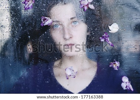 Closeup portrait of beautiful brunette woman behind the window with water drops on it and flowers