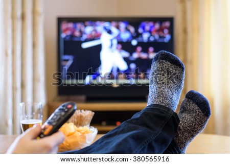 Television, TV watching (baseball match) with feet on table eating snacks and drinking beer - stock photo