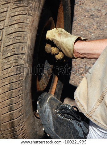 Man\'s hand bolts on a the spare tire after a blow out.  Sneaker clad foot holds pressure on bottom of tire.
