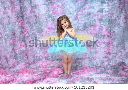 Ballerina wearing  aqua tutu and fairy wings stands in a room filled with pink and grey.  Her hands are folded against her cheek.