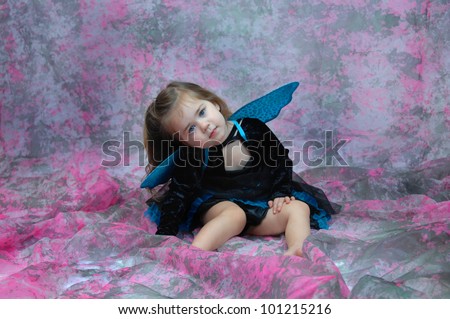 Adorable little girl is wearing a fairy costume in a room filled with pink and grey. Her eyes are vivid blue and match her wings and skirt.
