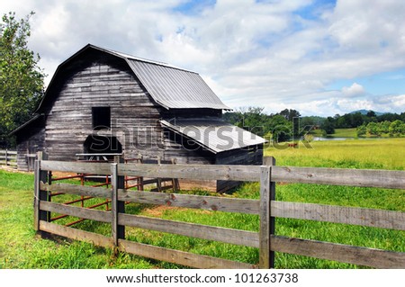 Horse grazes in the pasture close to a distant pond.  Barn and rustic wooden fence are both weathered and worn.  Blue sky and clouds make for perfect country scene.