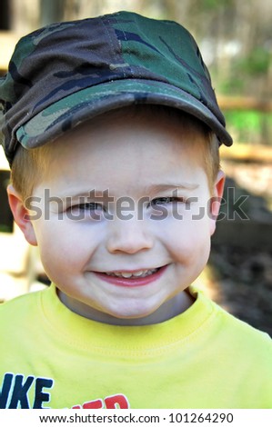 Beaming little boy is wearing a smile from ear to ear and a camouflaged hat.
