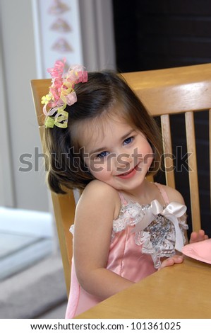 Little girl smiles happily at her ballerina birthday party.  She is sitting at a wooden table holding a pink paper plate.  She is wearing a pink costume.