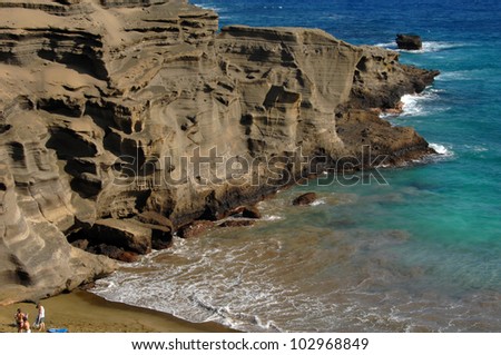 Three beach goers stand besides the sand stone cliffs on Green Sand Beach.  Their minute size in comparison to the cliffs give some scale to their tremendous size.  Aqua blue waters frame image.