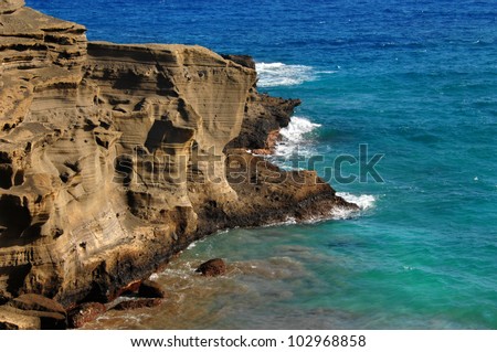 Cliff side of the Green Sand Beach, Big Island of Hawaii, wind and wave erosion is seen in the sand stone cliff face.  Aqua blue water and waves surround cliff.