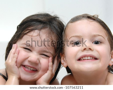 Two sisters laugh through bath time with wet heads and smiles.  Closeup shows one sister smiling and the other laughing.