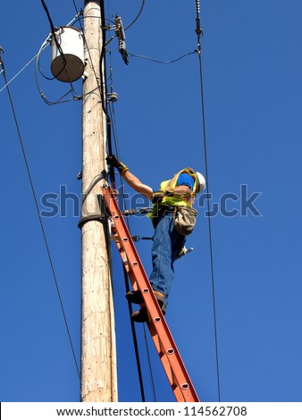Lineman climbs utility pole to fix problem.  He is standing on a ladder wearing a helmet and safety belt.  Blue sky frames him.