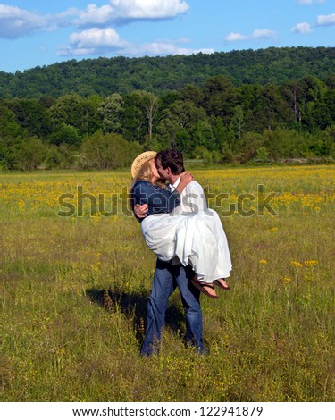 Young man carries wife across flower filled field.  He kisses her as she clings to his shoulders.  She is wearing a denim jacket and cowboy hat.