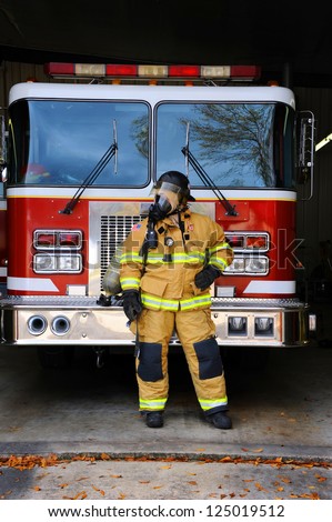 Woman firefighter stands in front of fire truck at fire station.  She is wearing bunking gear complete with gas mask and helmet.