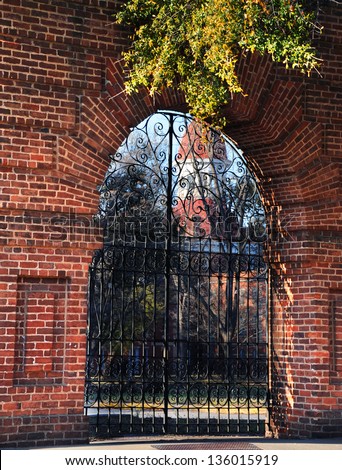 Derelict ruins of the Columbia State Mental Hospital in Columbia, South Carolina, sits behind locked, black metal gate.  Arched opening in brick wall gives peek to closed facility.