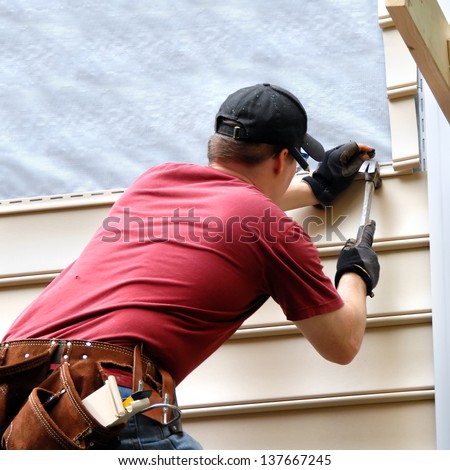 First Time Home Buyer Works To Install Siding On His New Home. He Is Hammering Into Place A Sheet Of Siding. He Has On A Red Shirt And Is Holding Hammer And Nail.