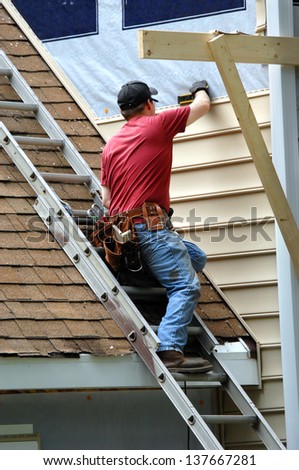 Young carpenter remodels aging home.  He is installing siding and is using a level to be sure installation is precise.  He is standing on a ladder on rooftop.