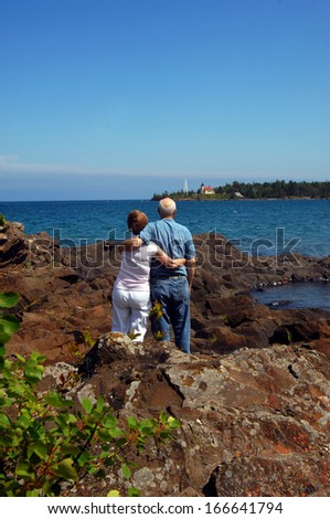 Affectionate, older couple stand on the shore of Lake Superior and admire the Copper Harbor Lighthouse in Upper Peninsula, Michigan.