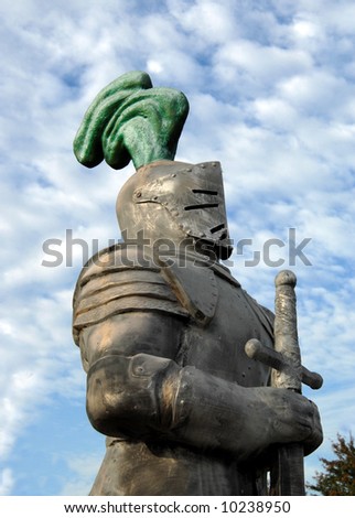 Knight stands tall against a blue sky with white clouds.  Plume of feathers on helmet are green.  He is holding a sword.