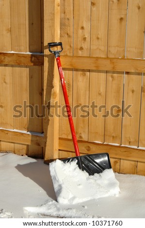 Snow shovel is propped against wooden security fence.  Handle is red and blade is black.  Shovel has a scoop full of fresh fallen snow.
