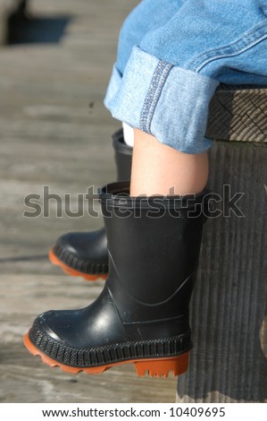 Small boy is wearing black rubber boots.  His denim overalls are rolled up.  Wooden dock and bench.  Sunshine.