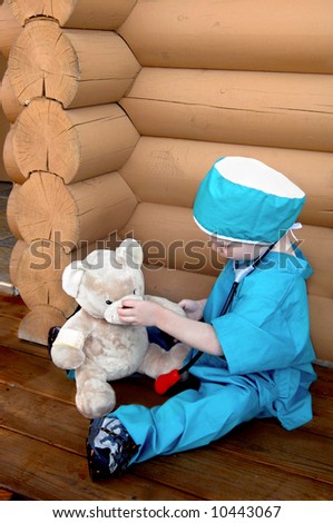 Young doctor works over his patient.  He has on a scrub suit and is wearing a stethoscope.  Teddy bear is his patient.  Log cabin in background.