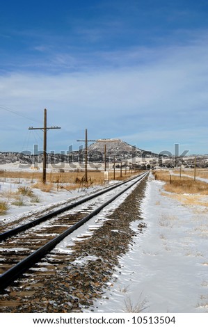 Railroad track disappears into the distance.  Winter scene with snow on tracks.  Mesa looms in distance.  Blue skies.