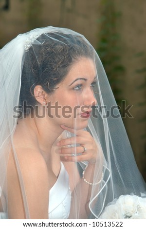 Beautiful young bride has veil over her head and face.  Her hand is under her chin and she is looking away.