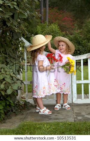 Two young sisters play in park besides a white wooden gate.  One girl lifts the brim of her sister's hat.