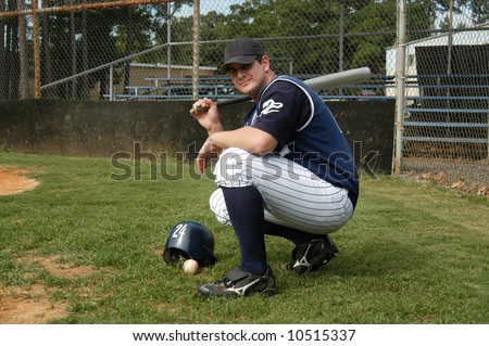 Young baseball player kneels on a grassy field.  He is holding a bat over his shoulder.  Helmet and ball sit on grass.