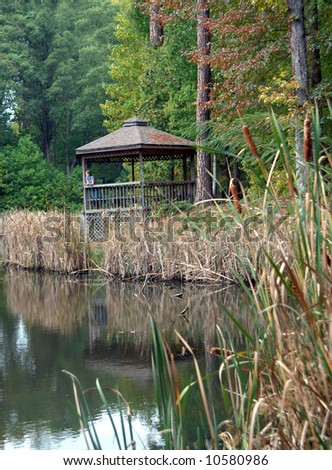Young woman leans on rail in wooden gazebo.  Gazebo faces lake.  Cattails surround lake.  Reflection is mirrored in surface of water.