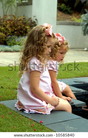 Two sisters sit on the side of a quiet pool.  They are dangling their feet in the water.  Long hair, bows and dresses.