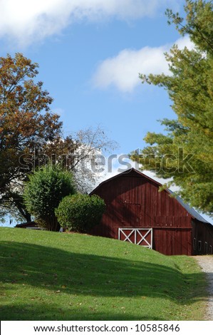 Red wooden barn sits on top of grassy slope.  Vivid blue sky and trees in background.