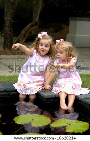 Two sisters play games while wetting their feet in waterlily pool.  Both have on pink dresses and bows.