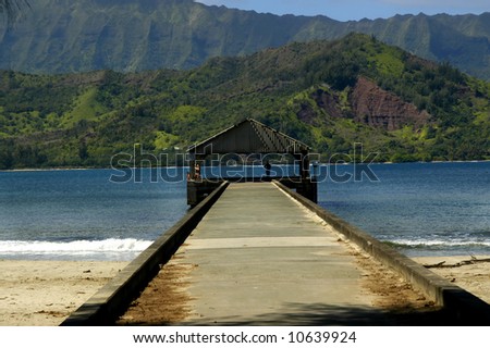 Hanalei Pier is framed by beautiful mountains and blue water. Concrete wharf leads out to a covered pavilion.