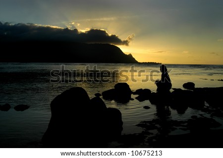 Woman is silhouetted as she sits on rocks in bay at Princeville Resort.  Sun is setting behind mountain and reflecting in water.